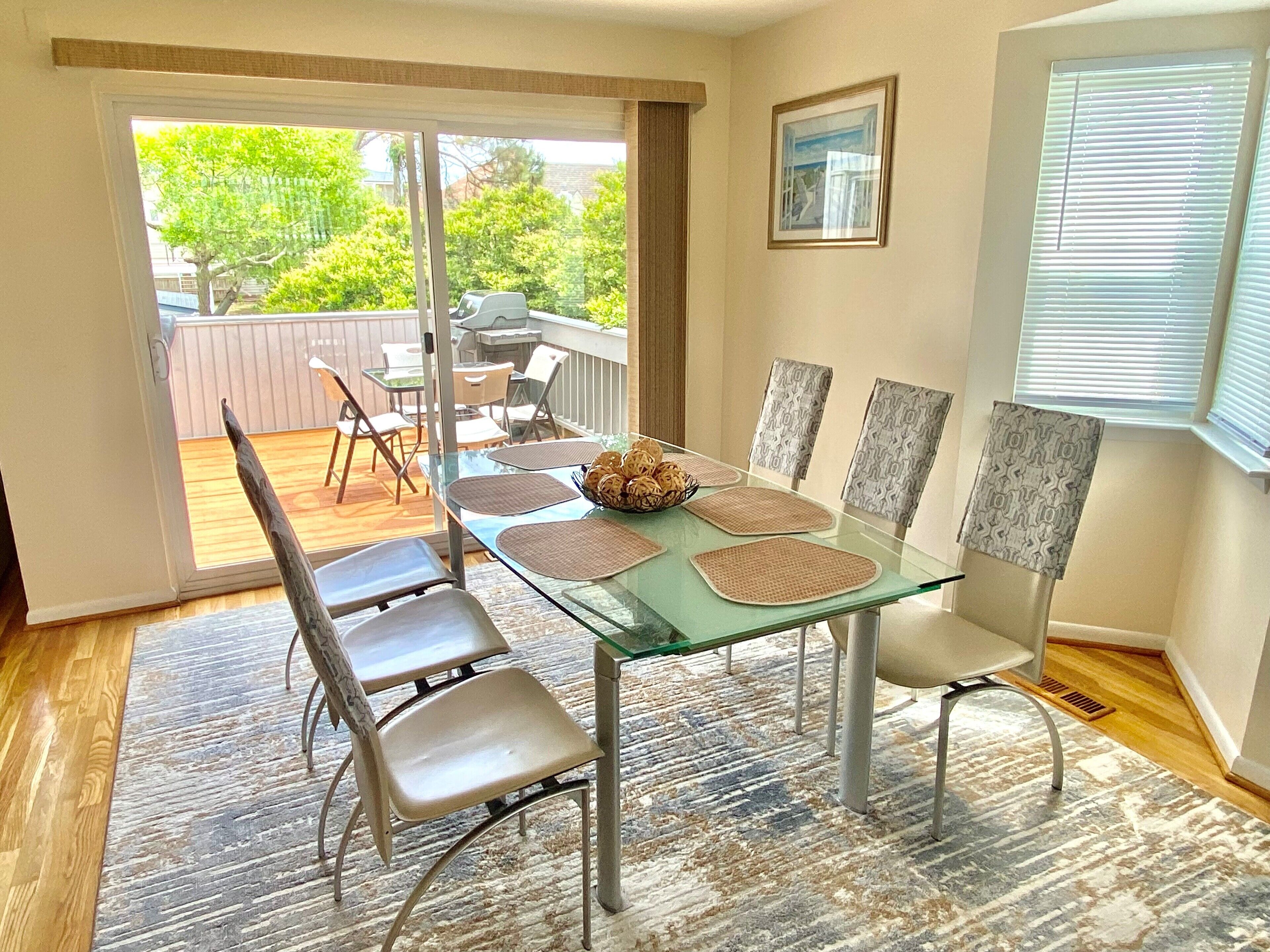 Een eetgedeelte met een glazen tafel en stoelen, uitzicht op de omgeving, en een raam met zonwering.