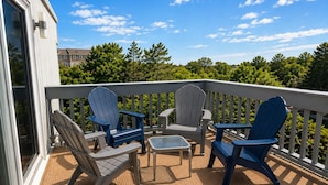 A balcony with Adirondack chairs, a small table, and a view of trees and buildings. - Sea La Vie: Ocean Side North End 3-Bedroom  - 2 min Walk to Beach (Virginia Beach)