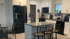 Una cocina moderna con gabinetes blancos, un refrigerador negro, un microondas y una cafetera. Hay una barra con taburetes y una mesa de comedor con sillas.
