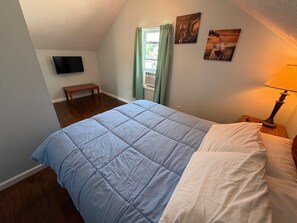 Een slaapkamer met een bed, nachtkastje, lamp, raam met gordijnen, een televisie aan de muur, en kunstwerken aan de muur.