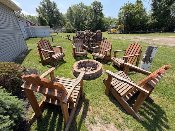 A backyard with a fire pit, wooden chairs, and a stack of firewood.