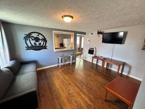 A living room with a sofa, a TV, a wooden floor, and a kitchen area in the background.