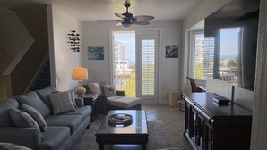 A living room with a grey sofa, a coffee table, a TV, and a ceiling fan. There is a door leading to a balcony with a view of buildings. - Luxury End-Unit Townhome: Private Elevator, Rooftop Hot Tub & Gulf Views (Clearwater)
