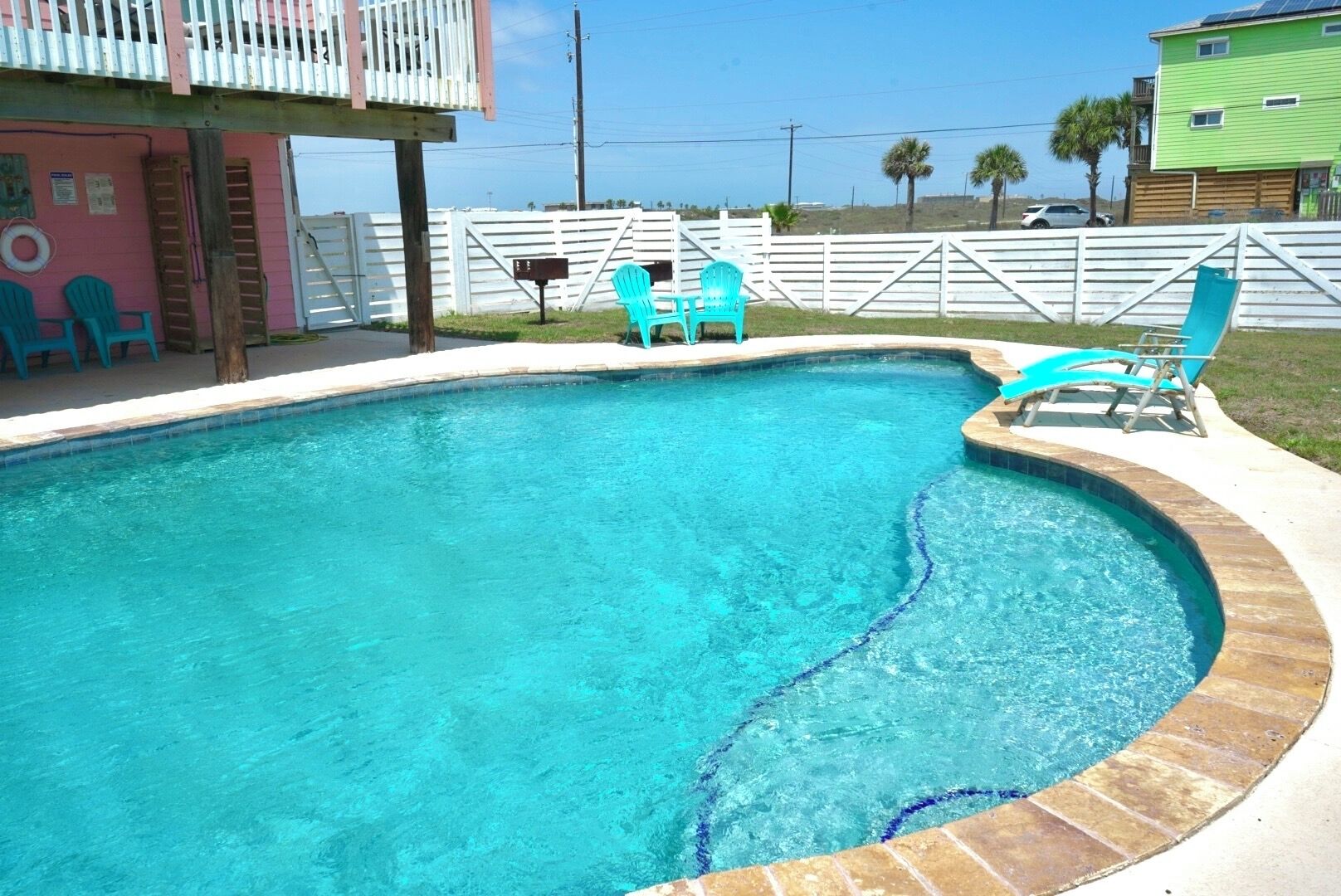 A swimming pool with a curved edge, surrounded by a white fence, and a small deck area with chairs.