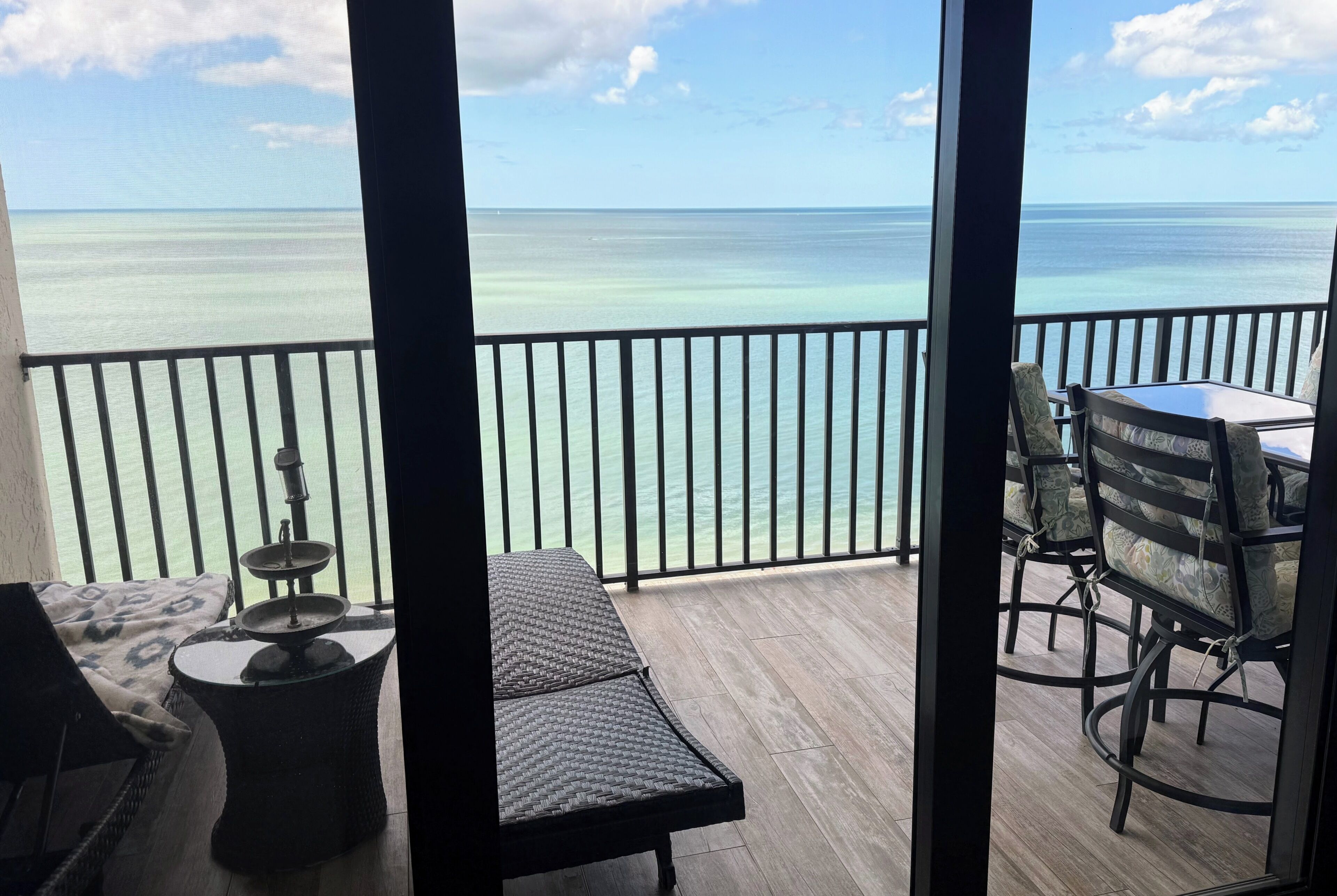 A balcony with a view of the ocean, a table, chairs, and a small round table with a lamp.