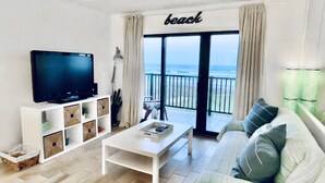 A living room with a television, a coffee table, a sofa, and a view of the beach through a large sliding door.