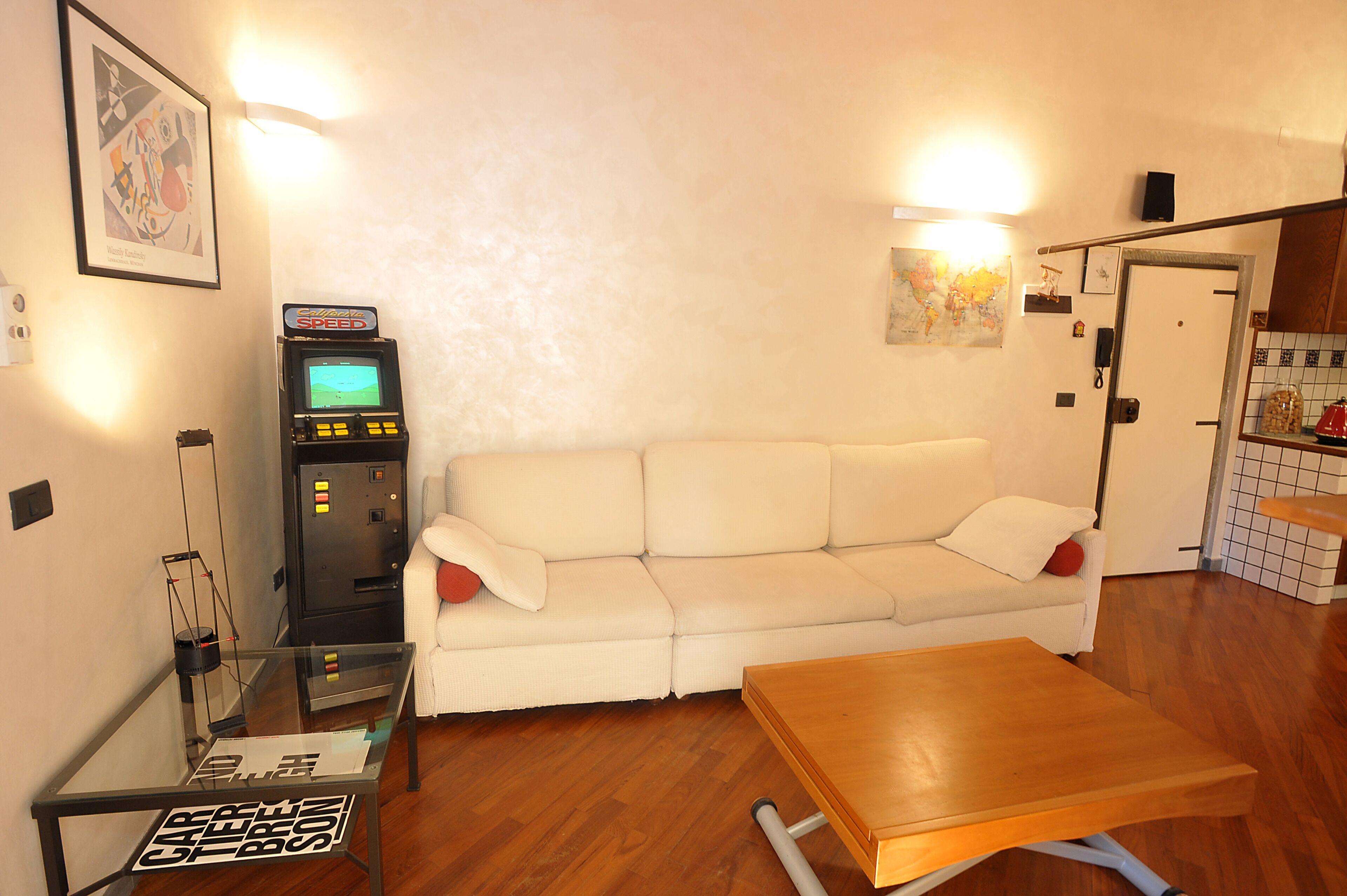 A room with a white sofa, a wooden table, a gaming machine, and a small glass table with a sign.