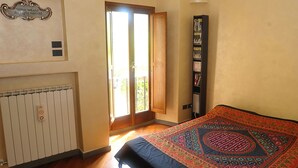 Un dormitori amb un llit gran, un radiador, una estanteria i una porta que dona a un balcó.