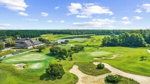 A golf course with a clubhouse, a pond, and a forested area in the background.