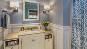 A bathroom with a beige vanity, a mirror, two wall-mounted lights, a towel rack, and a patterned shower curtain.