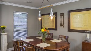 A dining room with a large wooden table, six chairs, a vase with flowers, and two hanging light fixtures.