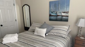 A neatly made bed with striped bedding, a painting of boats on the wall, a mirror, and a bedside table with a lamp.