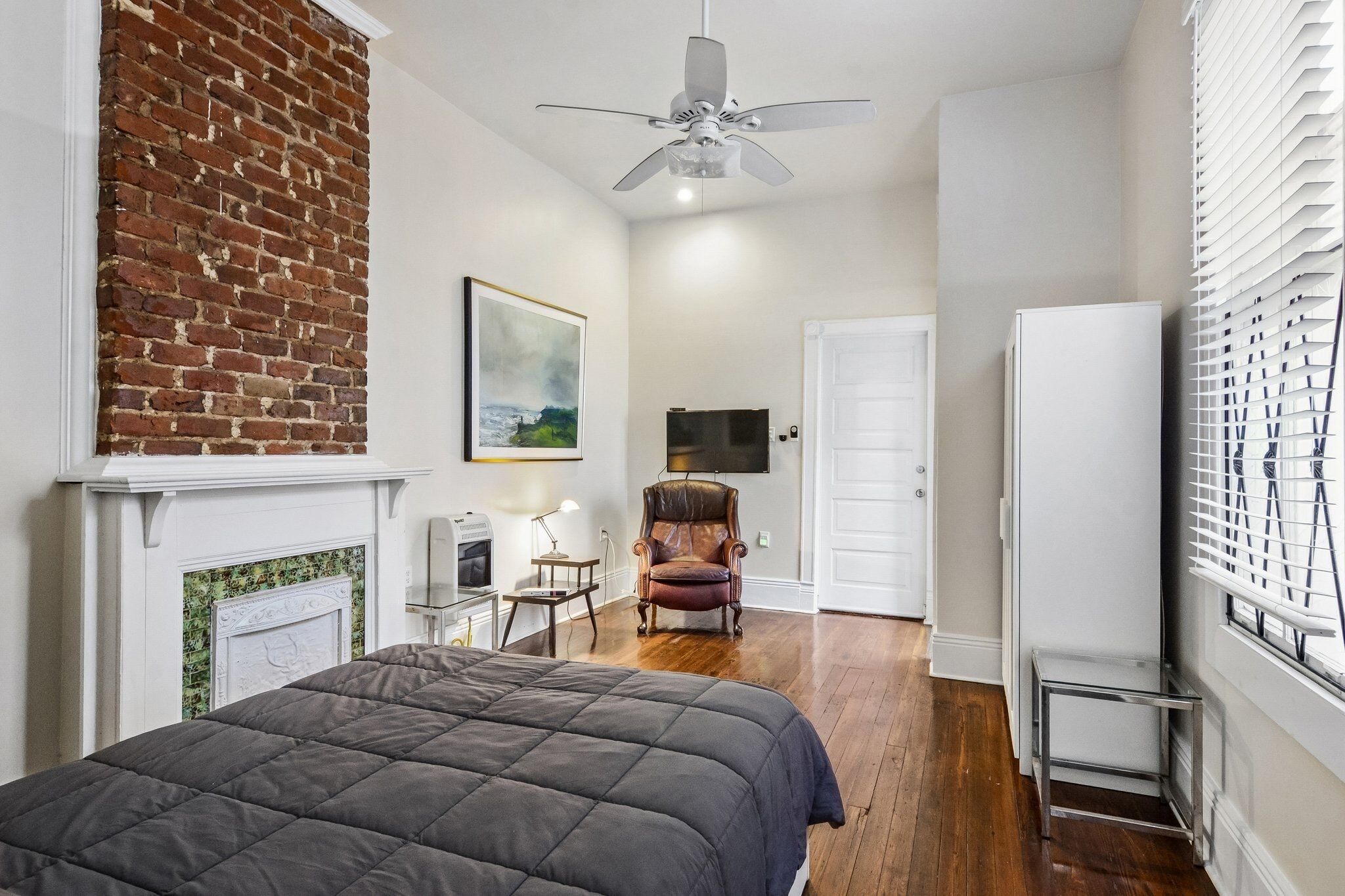 A bedroom with a bed, a fireplace, a brick wall, a ceiling fan, a television, a chair, and a door.