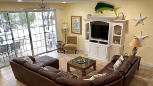 A living room with a brown leather sectional sofa, a glass coffee table, a white entertainment unit with a flat-screen TV, and a large window with a view of the outdoors.