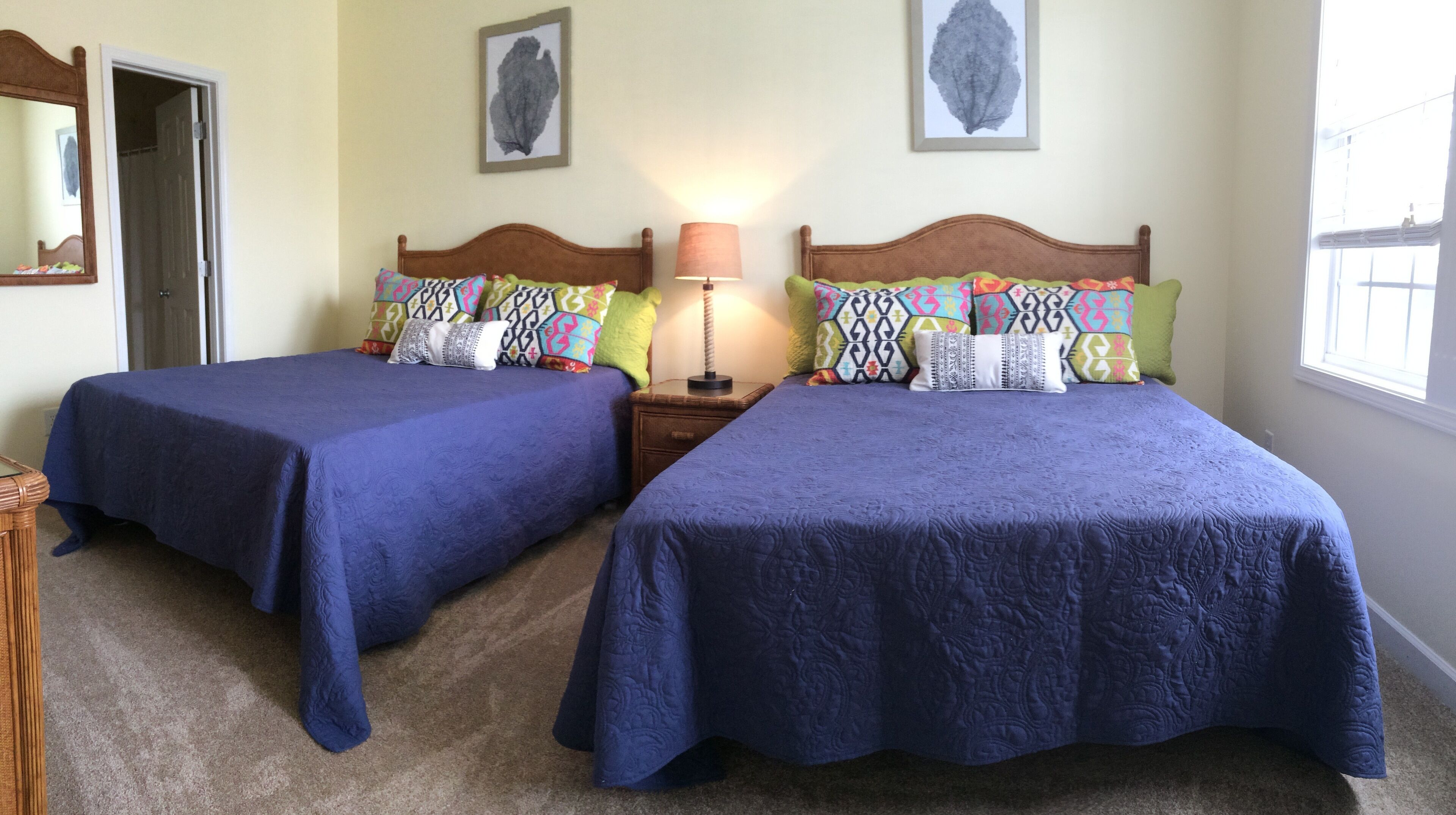 Une chambre avec deux lits, chacun recouvert d’une courtepointe bleue et agrémenté de coussins colorés. Une table de chevet avec une lampe se trouve entre les deux lits, et une porte est située sur le côté gauche.