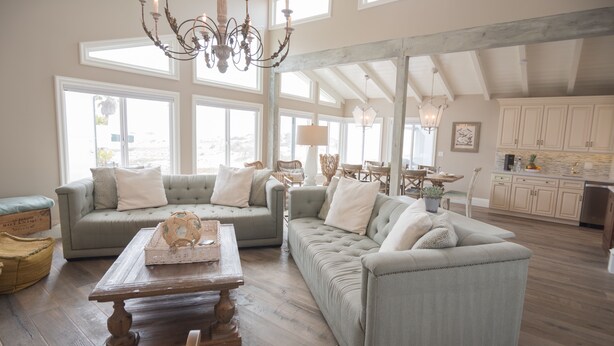 A spacious living room with two sofas, a wooden coffee table, and a chandelier. Large windows provide natural light, and the room features exposed wooden beams.