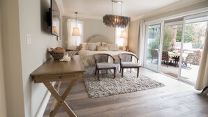 A bedroom with a bed, bedside tables, a desk, chairs, a chandelier, and a view through a sliding glass door to an outdoor patio.