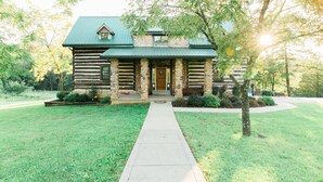 A log cabin with a stone porch, a green roof, and a pathway leading to the entrance. - Beautiful secluded Log Home - Nestled in the country! (Wright City)