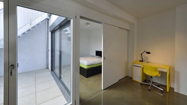 A modern bedroom with a bed, a desk, and a chair. There is a sliding door leading to a balcony. - Modern, Architect-Designed Penthouse Loft With 2 Outdoor Roof Terraces (Vienna)