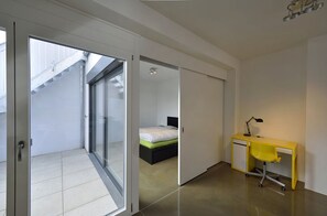 Ein modernes Schlafzimmer mit Bett, Schreibtisch und Stuhl. Eine Schiebetür führt zu einem Balkon.