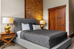 A hotel room with a large bed, two bedside lamps, a wooden nightstand, a brick accent wall, and a wooden door.
