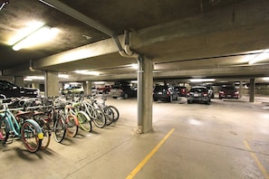 A multi-level underground parking garage with concrete pillars, fluorescent lighting, and a variety of parked bicycles and cars.