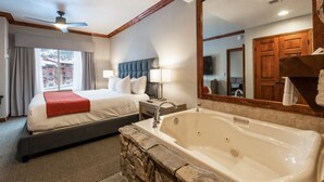 A hotel room with a large bed, a separate sitting area, a stone accent wall, a large bathtub, and a view of the outside.