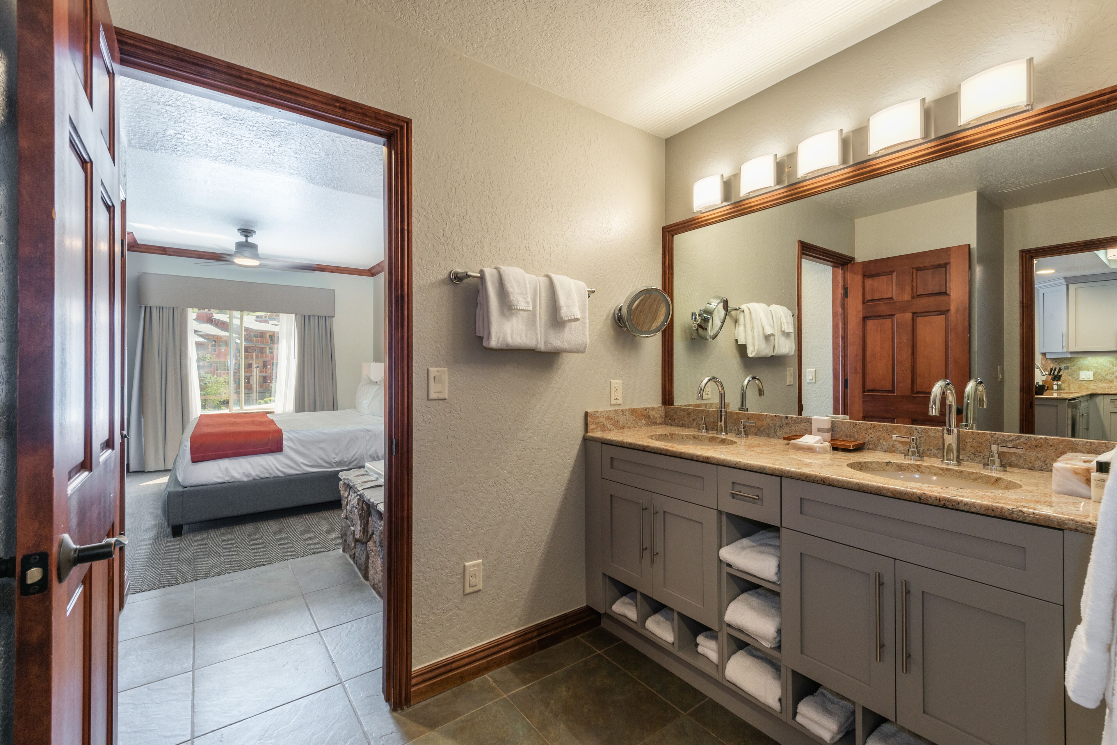A hotel bathroom with a large mirror, a sink, and a shower. There is a bed visible in the adjacent room.