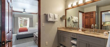 A hotel bathroom with a large mirror, a sink, and a shower. There is a bed visible in the adjacent room.