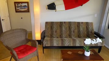 Een woonkamer met een rieten stoel, een bank, een bijzettafeltje met een plant, een surfplank en een ingelijst foto aan de muur.