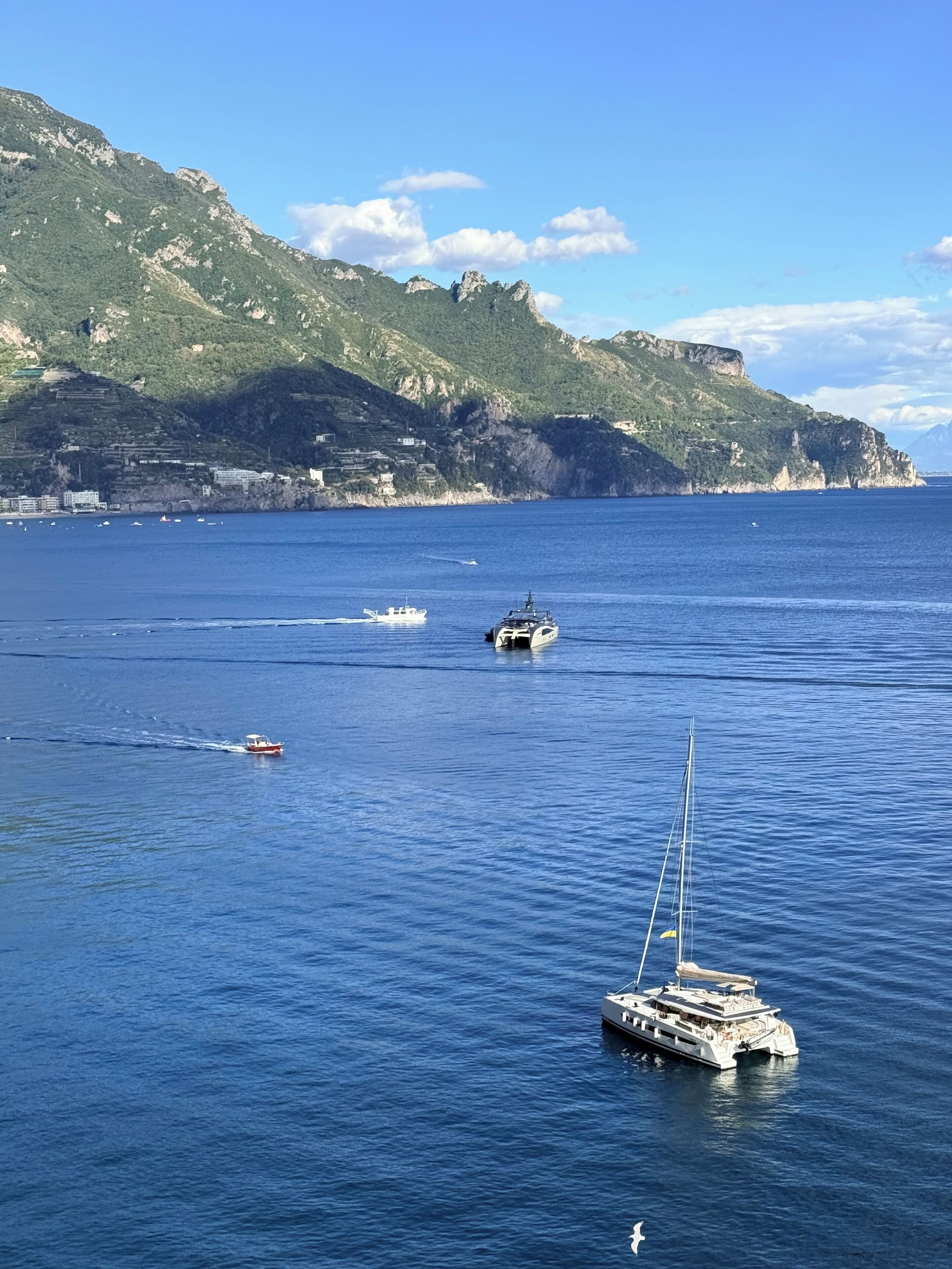 A coastal scene with boats on the water, a sailboat in the foreground, and a mountainous backdrop.