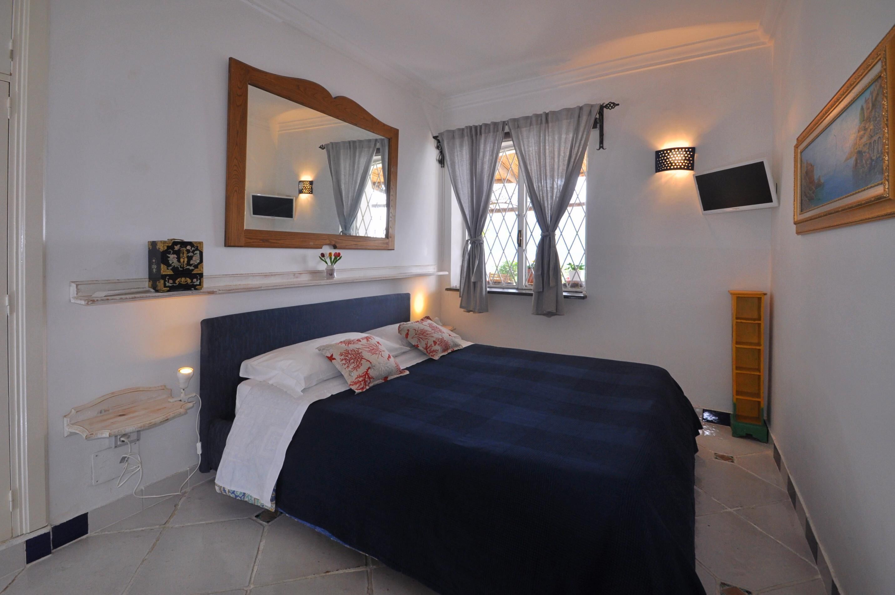 Une chambre à coucher comprenant un lit, un miroir, une étagère avec des objets décoratifs, un téléviseur fixé au mur et une fenêtre avec des rideaux.