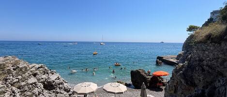 A rocky beach with clear blue water, several umbrellas, and boats in the distance.