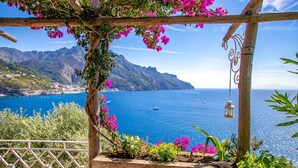 Eine hölzerne Pergola mit rosa Blüten, mit Blick auf eine Küstenlandschaft mit Bergen und dem Meer.