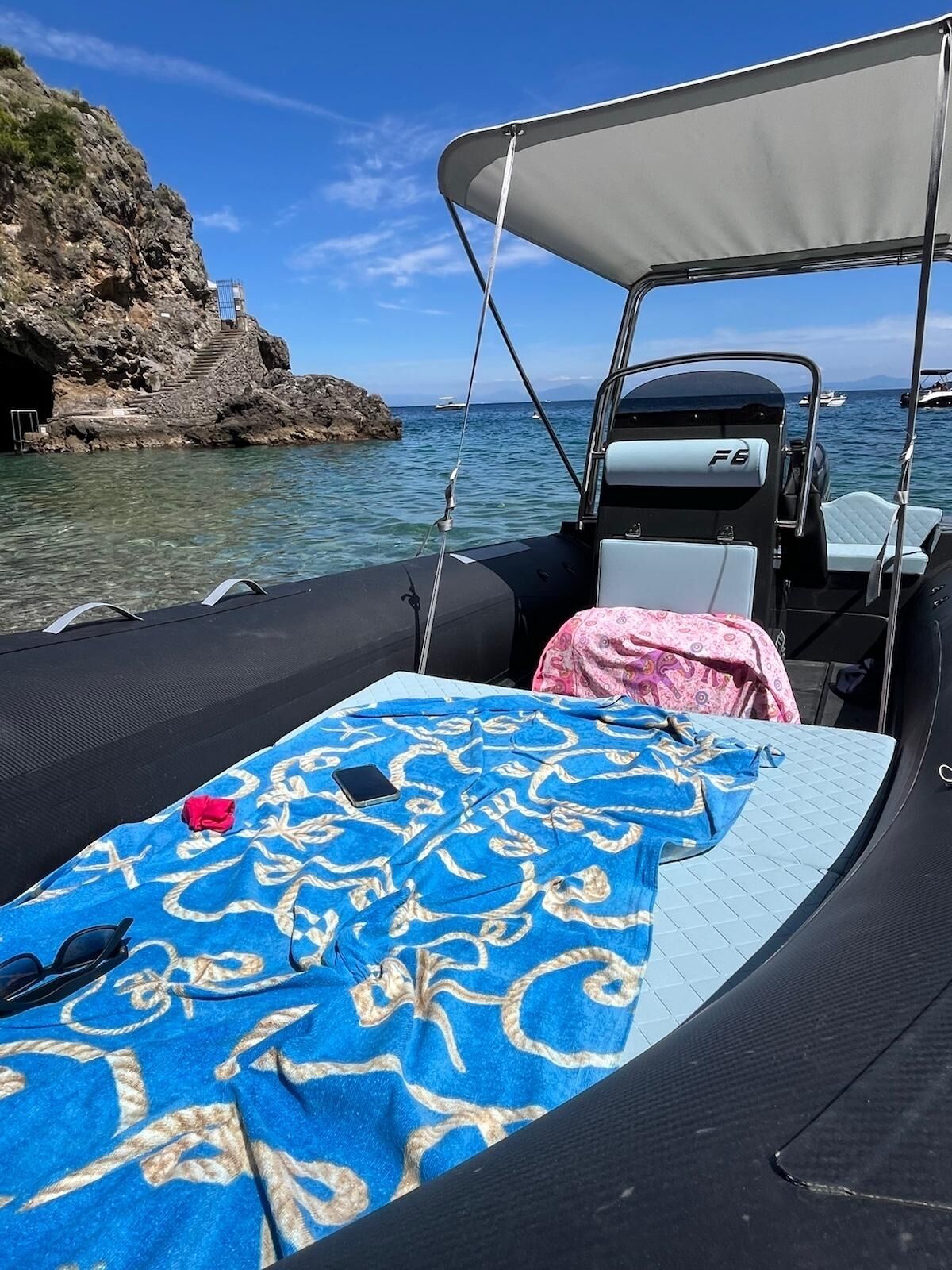 Ein Boot mit überdachtem Sitzbereich, einem kleinen Tisch mit blau gemustertem Tischtuch und einer felsigen Küstenlinie im Hintergrund.