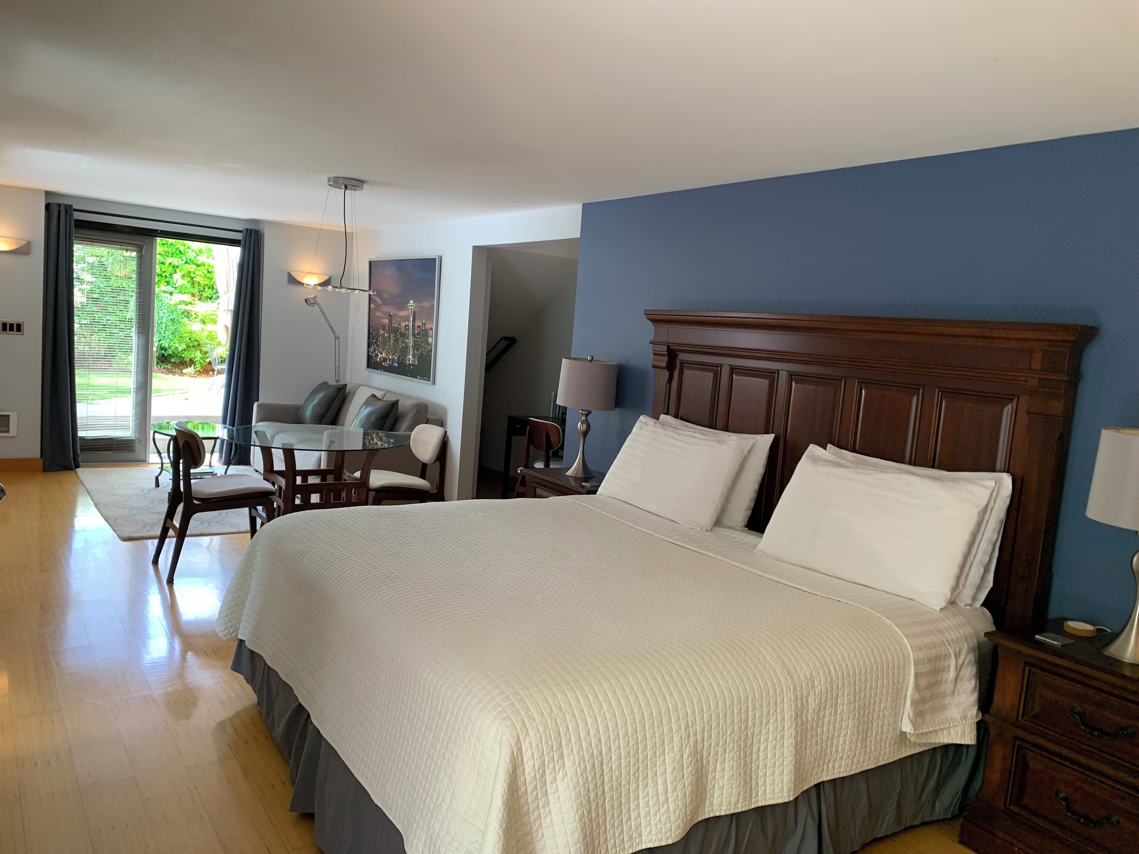 A bedroom with a large bed, a desk, and a chair. There is a window with curtains and a view of greenery outside.