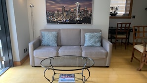 Sala de estar com sofá, mesa de centro de vidro e pintura de paisagem urbana na parede.