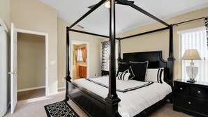 A four-poster canopy bed with a black frame, white bedding, and black and white pillows. There is a bedside table with a lamp and a black dresser with drawers. The room has beige walls and a carpeted floor. - The World Villa , South-Facing Pool & Spa , Free Wi-Fi , 3 Miles to Disney (KISSIMMEE)