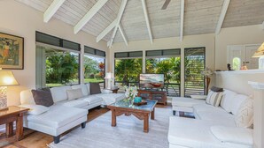 Un salon spacieux avec un plafond haut, de grandes fenêtres et une vue sur la végétation tropicale.