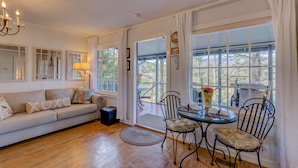 A cozy living room with a sofa, a round table, and chairs, a chandelier, and large windows with curtains.