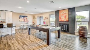 A modern living room with a pool table, bar stools, a fireplace, and a large window.
