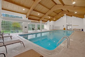 An indoor swimming pool with a wooden ceiling, clear glass windows, and a view of the outdoors.