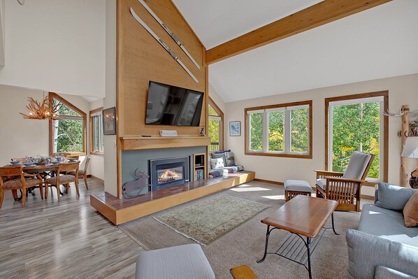A spacious living room with a fireplace, wooden beams, a TV mounted on the wall, and large windows offering a view of trees.