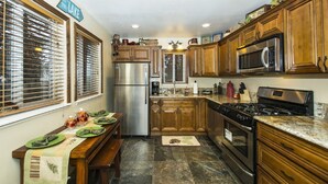 A compact kitchen with wooden cabinets, a stainless steel refrigerator, a microwave, and a stove. There is a dining table with green plates and a bench. - Gorgeous Lakeside hideout with views of the lake and mountains (Big Bear Lake)