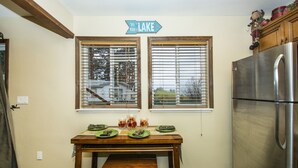 A kitchen with a stainless steel refrigerator, a wooden table with plates and glasses, and a window with blinds. - Gorgeous Lakeside hideout with views of the lake and mountains (Big Bear Lake)