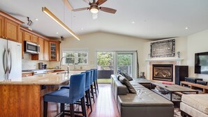 A modern kitchen with wooden cabinets, a bar area with blue barstools, a large grey sectional sofa, and a fireplace.