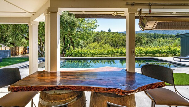 An outdoor patio with a wooden table and chairs, a pool, and a view of a lush garden and mountains.