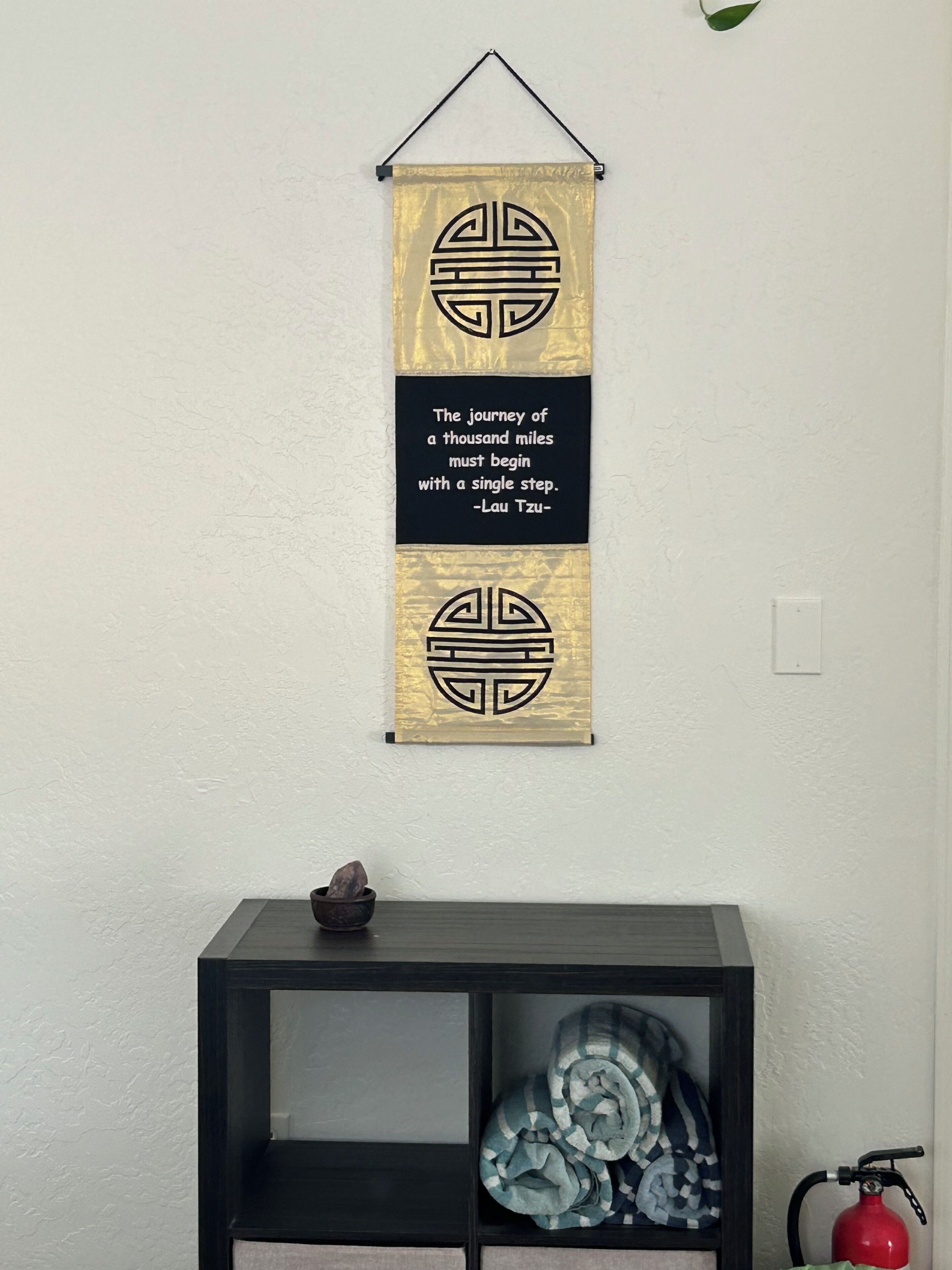 A wall with a hanging decorative panel featuring a quote and a geometric design. Below, there's a black console table with a bowl and a rolled-up blanket.