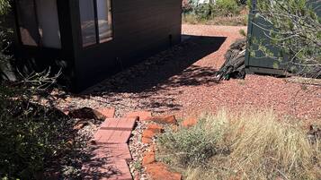 Una cabaña pequeña con techo inclinado, un camino de ladrillo que lleva a la entrada, y un paisaje circundante con árboles y arbustos.