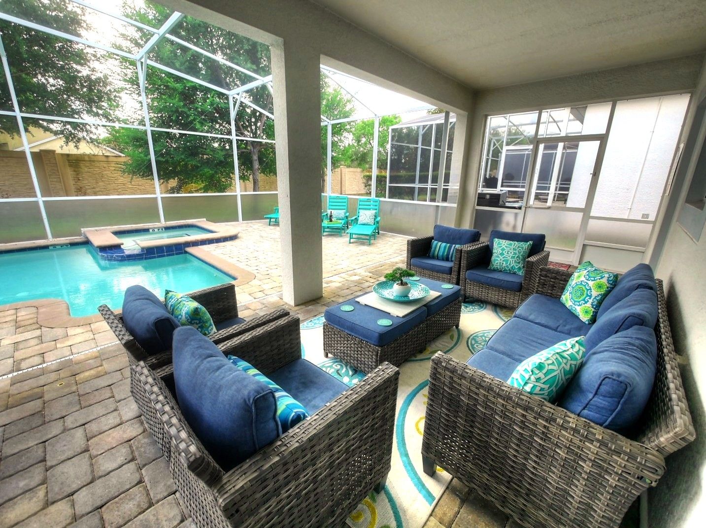 A covered outdoor area with a pool, wicker furniture, and a view of the surrounding greenery.